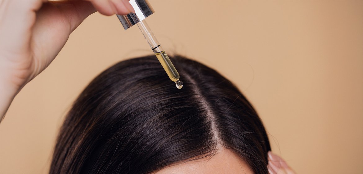 Nahaufnahme des Scheitels einer dunkelhaarigen Frau. Sie hält eine Pipette mit gelblicher Flüssigkeit über ihre Haare.